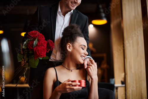 man holding red roses near happy african american woman with heart-shaped box on Valentines day