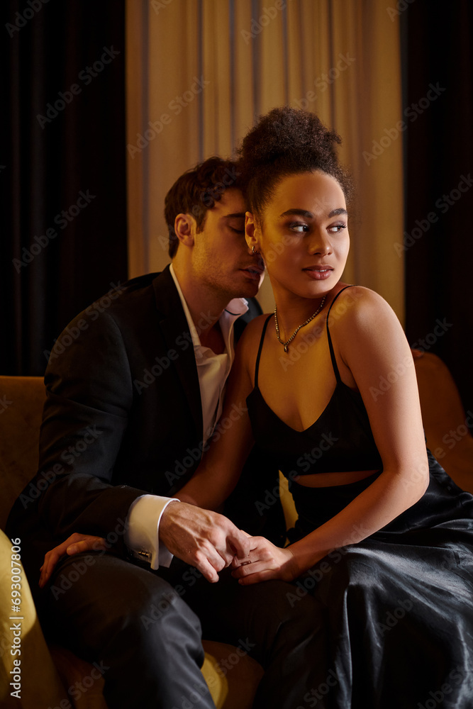 Obraz premium handsome man in suit seducing pretty african american woman in black dress and sitting on sofa