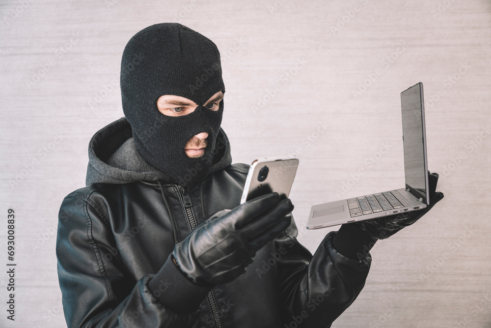 hacker thief uses a smartphone and laptop to steal money over the Internet. Criminal concept. Thief on a white background with a laptop hacker thief uses a smartphone and laptop to steal money over the Internet. Criminal concept. Thief on a white background with a laptop