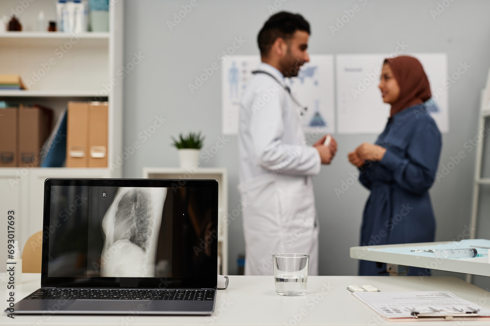 Medium shot of wireless laptop with X-ray scan of lungs on screen with talking doctor and patient in defocused background Medium shot of wireless laptop with X-ray scan of lungs on screen with talking doctor and patient in defocused background