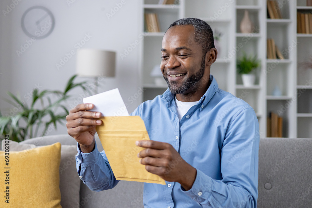 Senior joyful man sitting on sofa at home, satisfied smiling african american man reading mail message holding letter with good news in hands, inside living room of house. Senior joyful man sitting on sofa at home, satisfied smiling african american man reading mail message holding letter with good news in hands, inside living room of house.