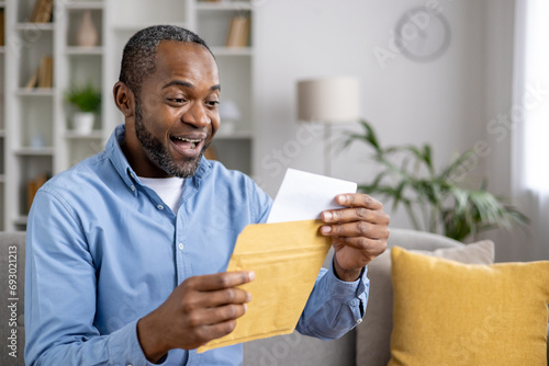 Senior joyful man sitting on sofa at home, satisfied smiling african american man reading mail message holding letter with good news in hands, inside living room of house. photo