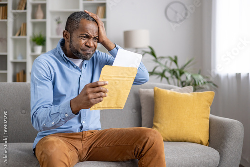 Fotografie Senior gray haired man upset received envelope mail message notification with bad news, sad displeased and disagreeable african american man sitting at home on sofa inside living room