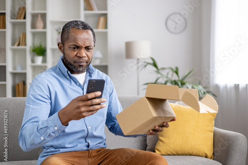 Fotografia Dissatisfied sad online buyer, man received wrong damaged parcel, african american man sitting at home on sofa in living room with cardboard box in hands and phone, writing negative review