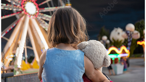 Uma menina de costas com um ursinho de pelúcia nos braços, olhando para um parque de diversões cheio de luzes coloridas. photo