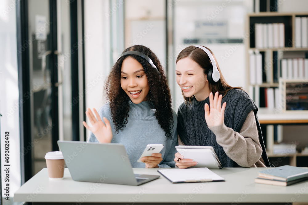 Two Attractive young female college students working on the school project using computer and tablet together, enjoy talking and headphones having a video chat. Two Attractive young female college students working on the school project using computer and tablet together, enjoy talking and headphones having a video chat.