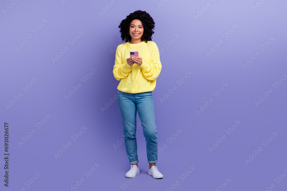 Full size photo of intelligent woman dressed yellow sweater denim pants texting message on smartphone isolated on violet color background Full size photo of intelligent woman dressed yellow sweater denim pants texting message on smartphone isolated on violet color background