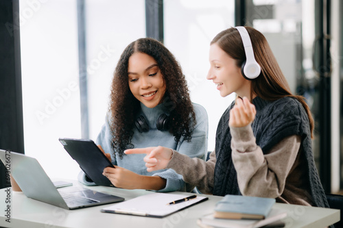 Two Attractive young female college students working on the school project using computer and tablet together, enjoy talking and headphones having a video chat.