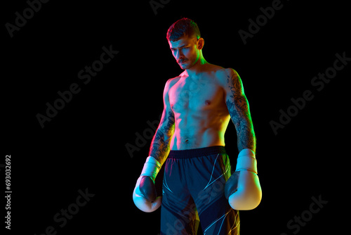 Young man with muscular strong shirtless body, boxing athlete standing isolated over black background in neon light. Concept of professional sport, combat sport, martial arts, strength © master1305