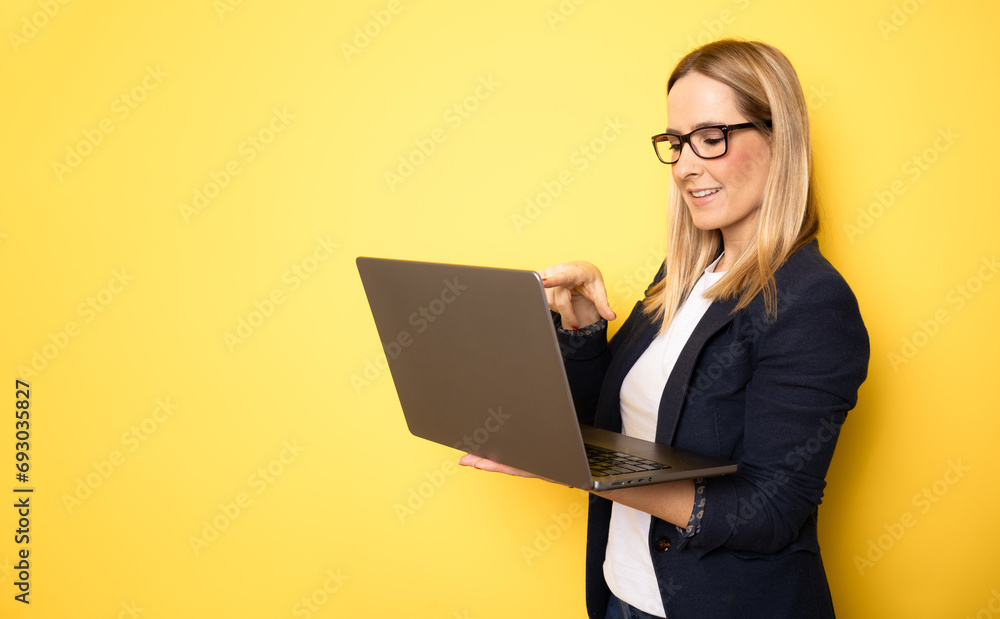 Smiling cheerful funny beautiful attractive young woman 40s wearing basic casual jacket standing working on laptop pc computer isolated on yellow colour background studio portrait Smiling cheerful funny beautiful attractive young woman 40s wearing basic casual jacket standing working on laptop pc computer isolated on yellow colour background studio portrait