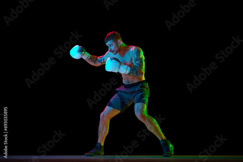 Full-length of shirtless young man with muscular strong body, boxer in motion, training isolated on black background in neon light. Concept of professional sport, combat sport, martial arts, strength