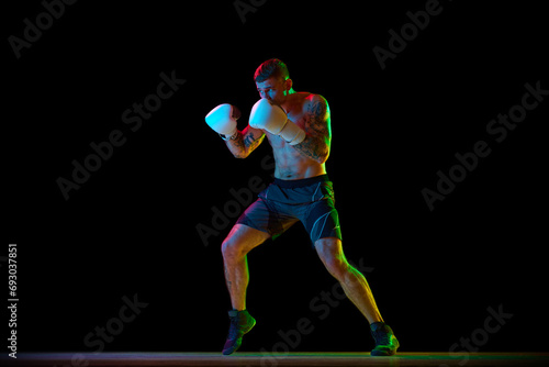 Fotografía Full-length of shirtless young man with muscular strong body, boxer in motion, training isolated on black background in neon light