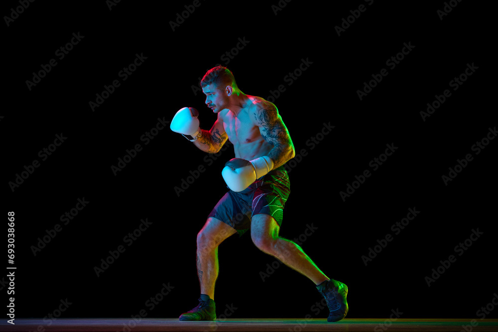 Full-length image of young athletic man, muscular, shirtless boxer in motion, fighting, practicing hooks isolated over black background in neon. Concept of sport, combat sport, martial arts, strength