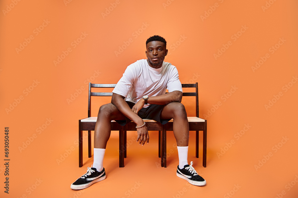 appealing young african american man in urban outfit sitting on bench on orange backdrop, fashion appealing young african american man in urban outfit sitting on bench on orange backdrop, fashion