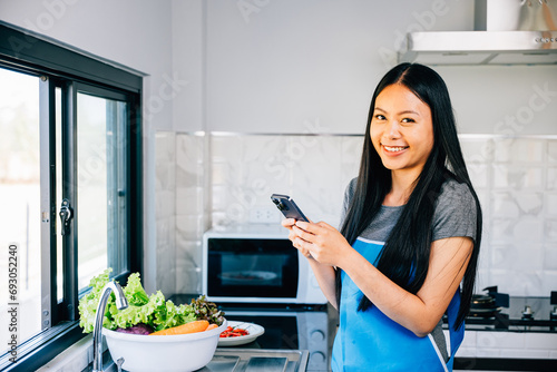 In a cozy kitchen an Asian woman cooks vegetables viewing a cooking tutorial on her smartphone. Smiling and inspecting fruits she merges modern tech with culinary exploration.