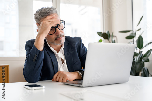 Thoughtful male office employee using laptop indoors, looking at laptop screen with puzzled face expression, businessman solving tasks, having problems with finance