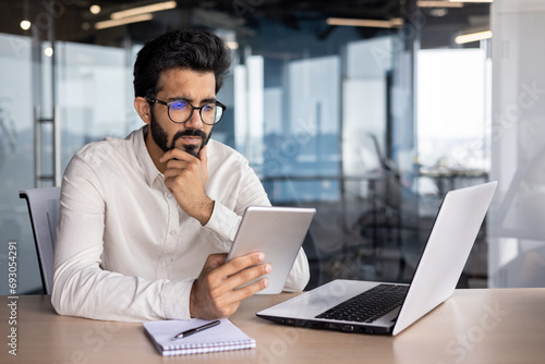 Concentrated and serious young Indian man working in office at desk with tablet photo