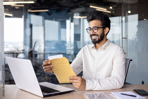 A smiling young man sits in the office at the table with a laptop and opens an envelope with a letter, reads good news