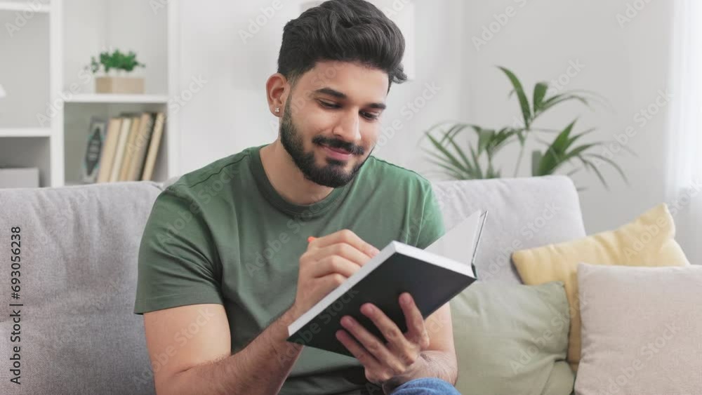 Positive bearded guy dressed in casual clothes sitting on comfy grey couch and writing with pen in notebook. Hindu young man taking notes during daydreaming at cozy modern apartment. Positive bearded guy dressed in casual clothes sitting on comfy grey couch and writing with pen in notebook. Hindu young man taking notes during daydreaming at cozy modern apartment.