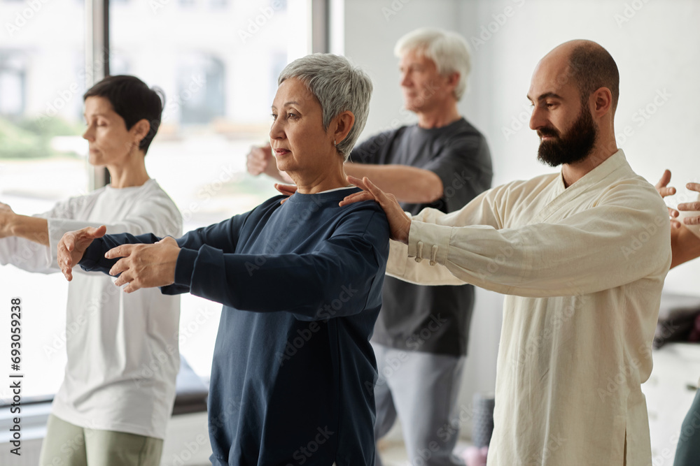 Obraz premium Side view of bearded male trainer correcting shoulder position of gray-haired senior woman during qigong exercise