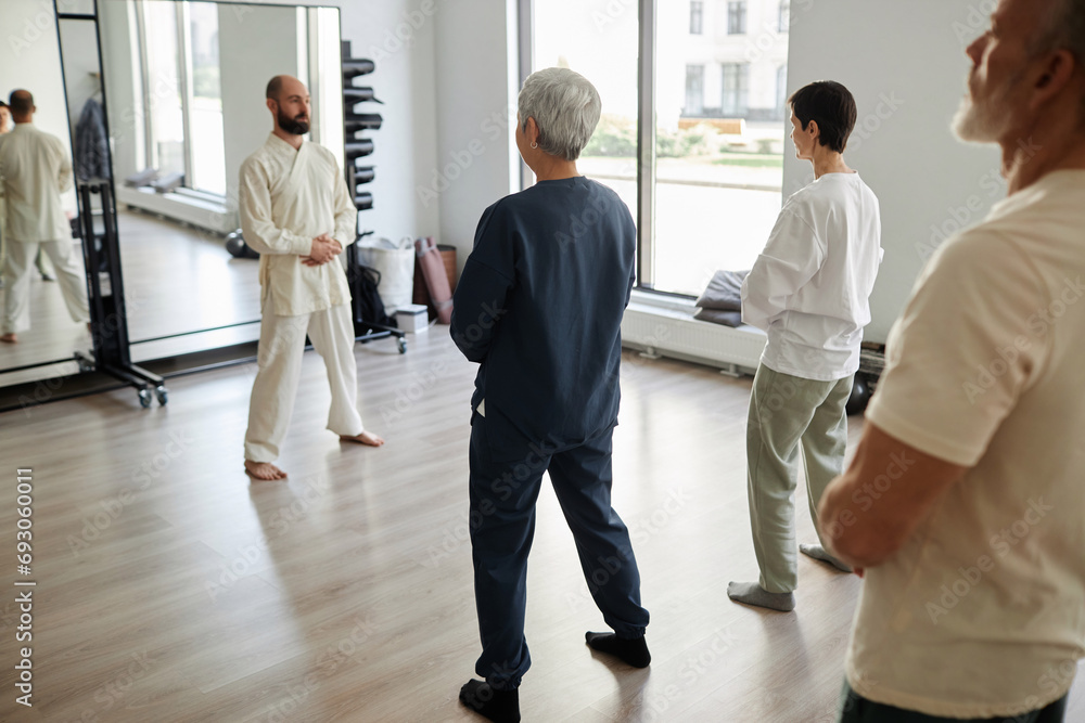 Fototapeta premium Back view at group of senior people standing in gym and meditating while qigong instructor observing