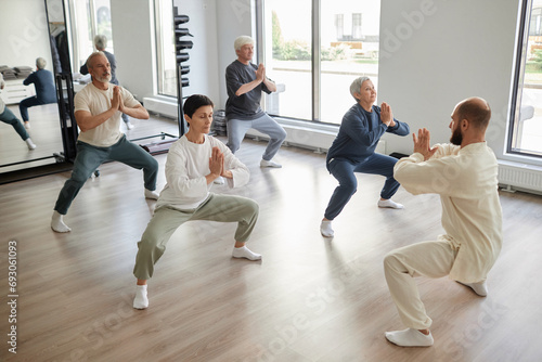 Wide angle view of elderly people and qigong instructor in gym standing in half-squat holding hands in prayer pose photo