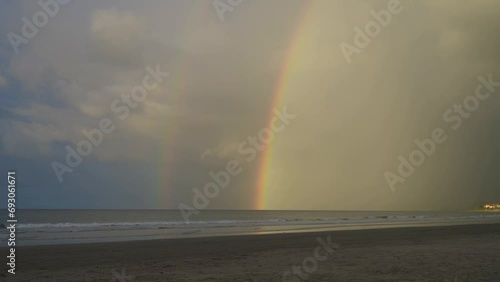 A double rainbow over a tropical beach divides the sky into two parts: a dark one on the right and a blue one with white clouds on the left. The ocean waves roll towards the shore, foaming.  Malaysia