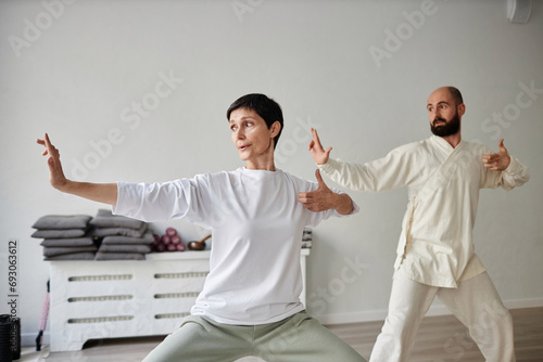 Wide shot of dark-haired adult woman and trainer practicing archer pose during qigong class in gym