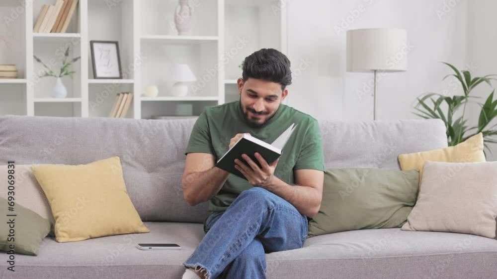 Excited indian guy sitting on comfy grey couch and pointing with finger up while reading book. Inspired bearded man satisfied with new idea or problem solution while doing hobby. Excited indian guy sitting on comfy grey couch and pointing with finger up while reading book. Inspired bearded man satisfied with new idea or problem solution while doing hobby.