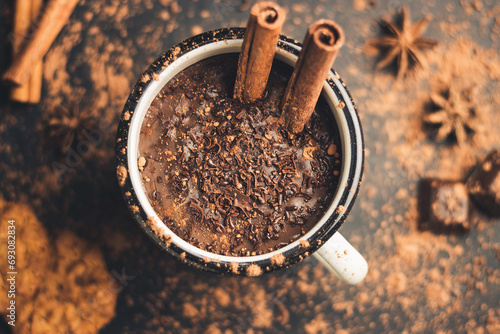 Homemade spicy hot chocolate drink with cinnamon stick, star anise, grated chocolate in enamel mug on dark background with cookies, cacao powder and chocolate pieces, top view photo