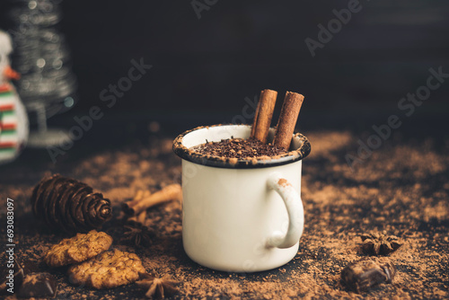 Homemade spicy hot chocolate drink with cinnamon stick, star anise, grated chocolate in enamel mug on dark background with cookies, cacao powder and chocolate pieces