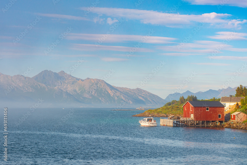 Beautiful seascape with low clouds or fog in Norway. View of the picturesque coast with the mountains, traditional red fisherman's cabins rorbu and a boat. Travel Scandinavian background