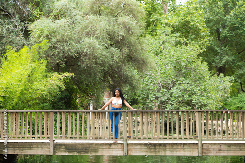 Young and beautiful brunette woman and latin, leaning on the railing of the wooden bridge over the duck and fish pond in the park. The woman is smiling and happy and enjoying the day.
