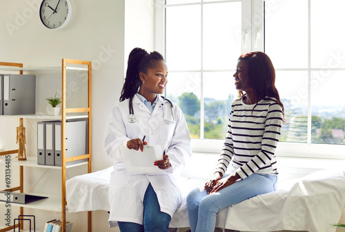Happy African American female doctor and patient discussing treatment. Young woman physician in white medical coat sitting on examination bed with young girl, talking about her therapy, giving advice photo