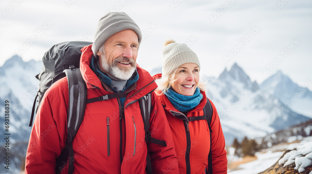 Two hikers (elderly couple) in winter gear enjoy a cold, adventurous hike against a snowy mountain backdrop. The image showcases the beauty of nature and the thrill of exploration. 