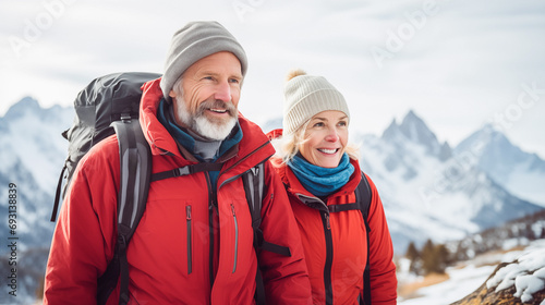 Two hikers (elderly couple) in winter gear enjoy a cold, adventurous hike against a snowy mountain backdrop. The image showcases the beauty of nature and the thrill of exploration. 