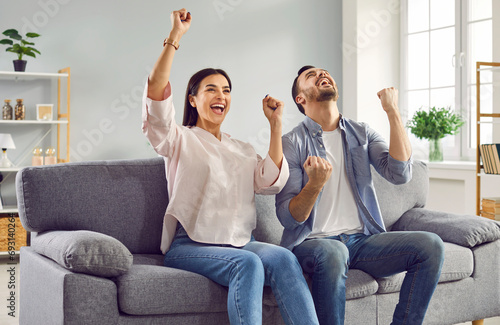 Happy young family couple raising hands on couch at home. Excited husband and wife celebrating goal achievement, victory of their favorite team, lottery win or moving in new house photo