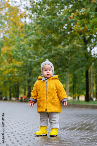 Cute baby toddler girl in a beautiful coat and yellow rubber boots walks in the park after the rain