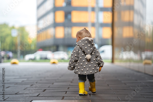 A cute stylish two-year-old toddler girl kid in a gray coat and yellow boots walks outdoors in the city in autumn season 