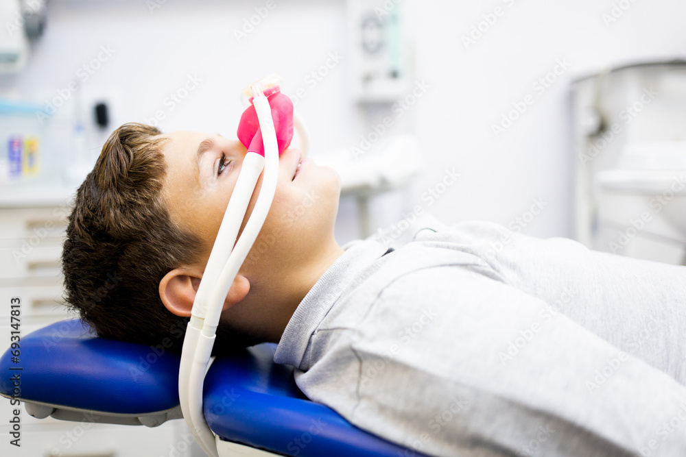 Fear of the dentist! A little boy sits in a dentist's office wearing a nasal mask breathing nitrous oxide to relax.Concept of feeling relaxed with laughing gas.Anxiety about visiting a dentist. - obrazy, fototapety, plakaty 