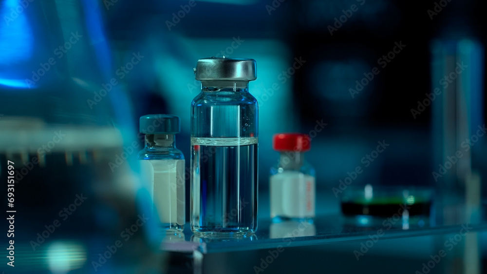 Close up of glass vials with clear liquid and petri dish on table in research lab or hospital. Vials with clear liquid on blurred background. Concept of laboratory research, health care. Close up of glass vials with clear liquid and petri dish on table in research lab or hospital. Vials with clear liquid on blurred background. Concept of laboratory research, health care.