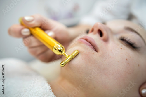 woman receive pulse face massaging at spa, vibrating golden facial massager on female face photo
