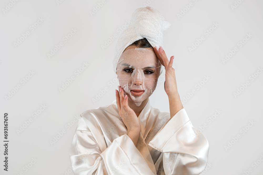 Young woman in white towel applies white cloth moisturizing mask isolated on white Young woman in white towel applies white cloth moisturizing mask isolated on white