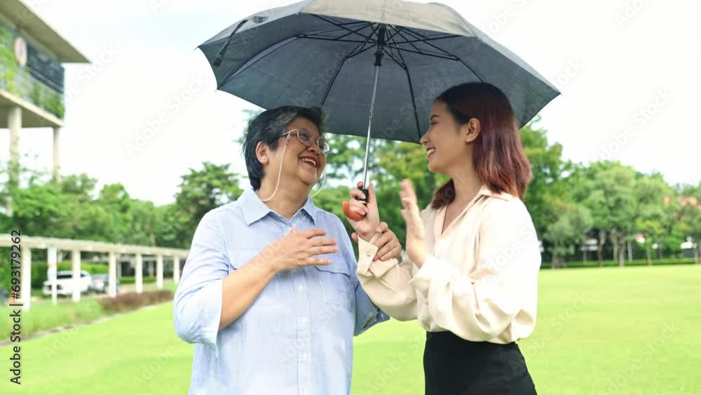 Elderly mother and her beautiful asian daughter walk together in the garden, holding up umbrella sun shade outdoors to protect their skin heatstroke from the sun and the heat of the hot weather. Elderly mother and her beautiful asian daughter walk together in the garden, holding up umbrella sun shade outdoors to protect their skin heatstroke from the sun and the heat of the hot weather.