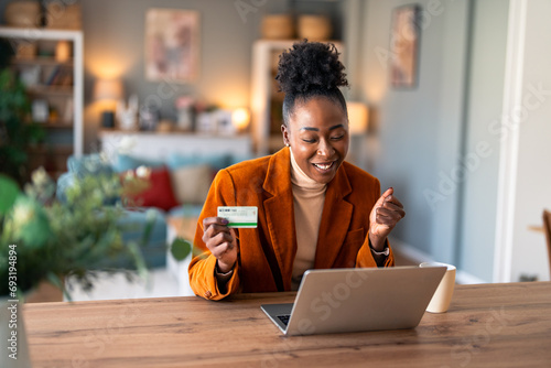Happy young female shopper holding credit card, using laptop doing online banking transaction while sitting at desk in home office during the day. photo
