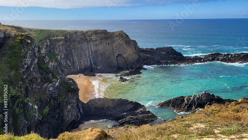 Praia de Almograve beach with ocean waves, cliffs and stones, wet golden sand and green vegetation at wild Rota Vicentina coast, Odemira, Portugal