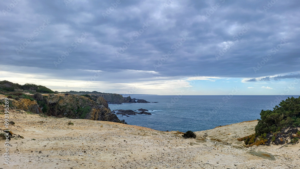 Zambujeira do Mar over the sea shore with ocean waves, cliffs and sand dunes covered by green vegetation red leaves of sour fig, sunny day, clear blue sky. Rota Vicentina coast, Portugal. Zambujeira do Mar over the sea shore with ocean waves, cliffs and sand dunes covered by green vegetation red leaves of sour fig, sunny day, clear blue sky. Rota Vicentina coast, Portugal.
