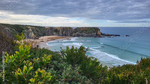 Zambujeira do Mar over the sea shore with ocean waves, cliffs and sand dunes covered by green vegetation red leaves of sour fig, sunny day, clear blue sky. Rota Vicentina coast, Portugal. photo