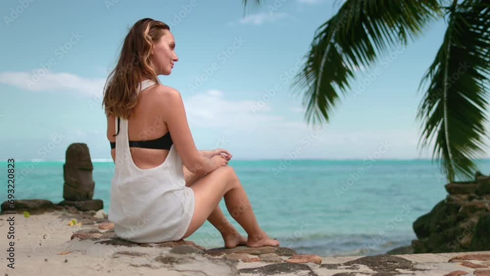 Woman relax on sea coast beach under palm tree. Barefoot girl in white shirt sit on shore enjoy exotic island landscape. Outdoor lifestyle travel, summer holiday vacation. Tropical scene. Slow motion Woman relax on sea coast beach under palm tree. Barefoot girl in white shirt sit on shore enjoy exotic island landscape. Outdoor lifestyle travel, summer holiday vacation. Tropical scene. Slow motion