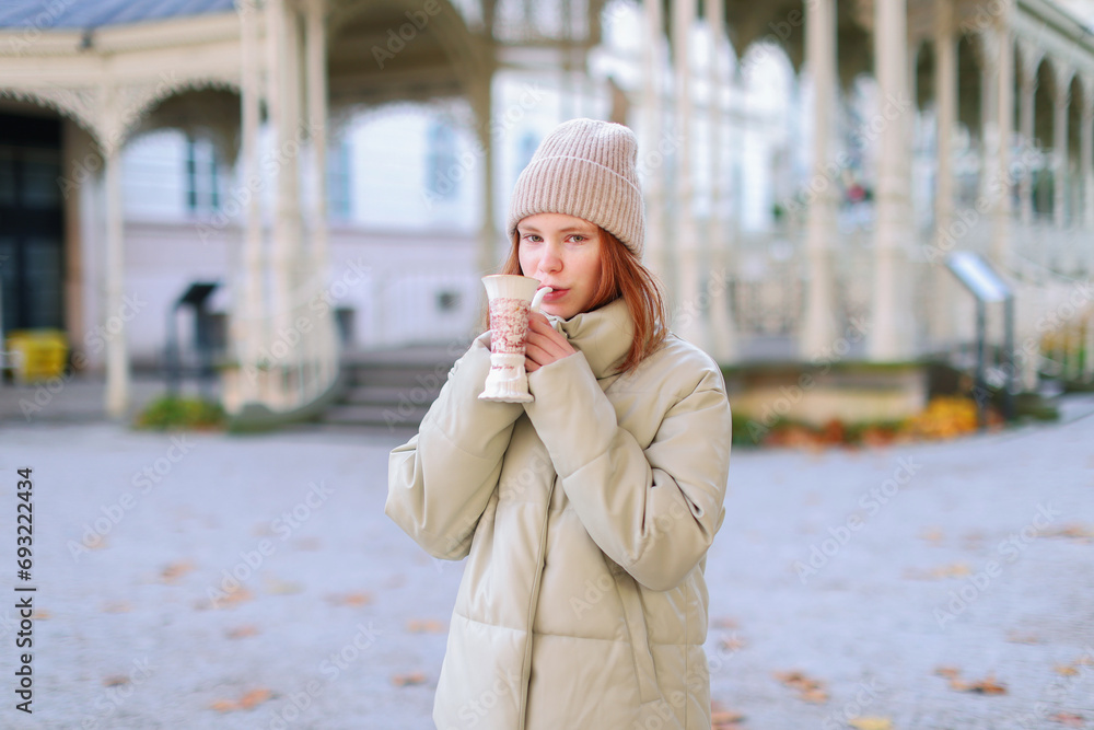 Fototapeta premium Young girl drinking from cup with therapeutic mineral water at a natural hot spring in Karlovy Vary